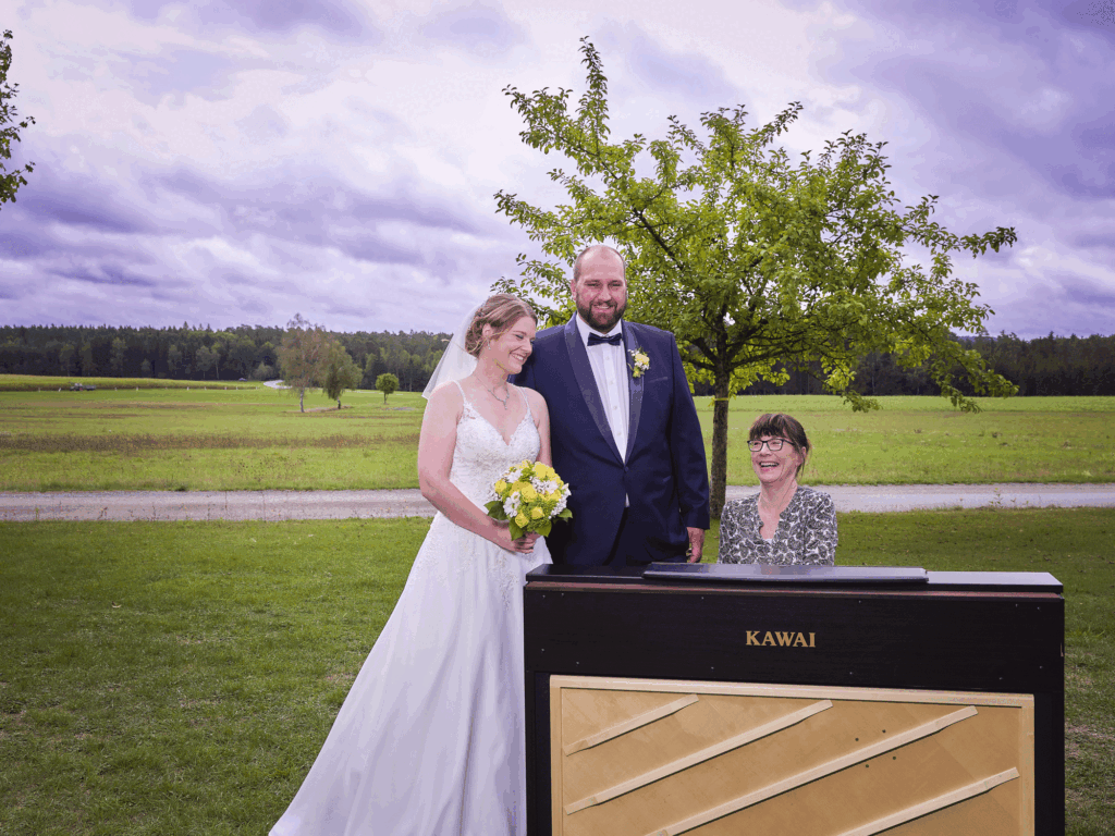 Bild in freier Natur. Mutter sitzt am Klavier. Das Brautpaar steht daneben.,