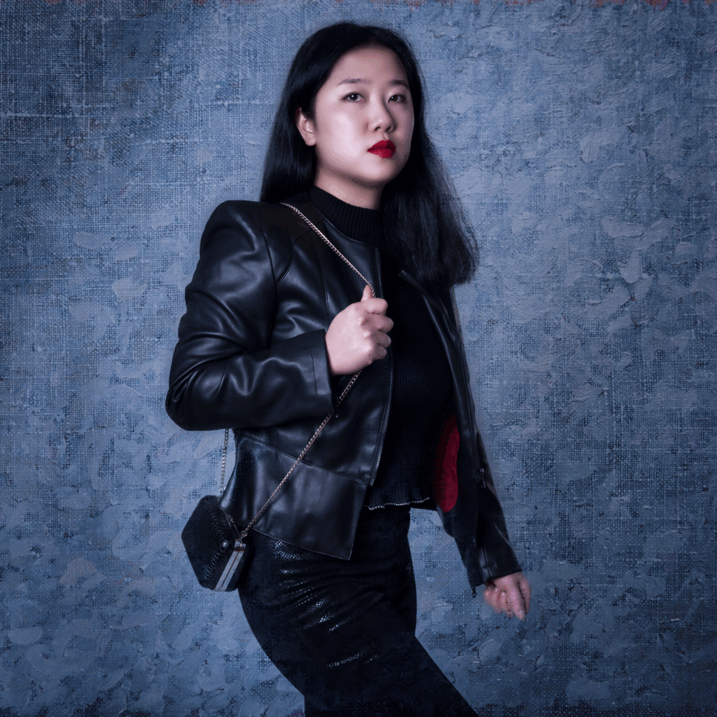 Halbkörperporträt einer asiatischen Frau mit langen, dunklen Haaren. Sie trägt eine schwarze Lederjacke vor einem blauen Hintergrund. Die roten Lippen geben Attraktivität.