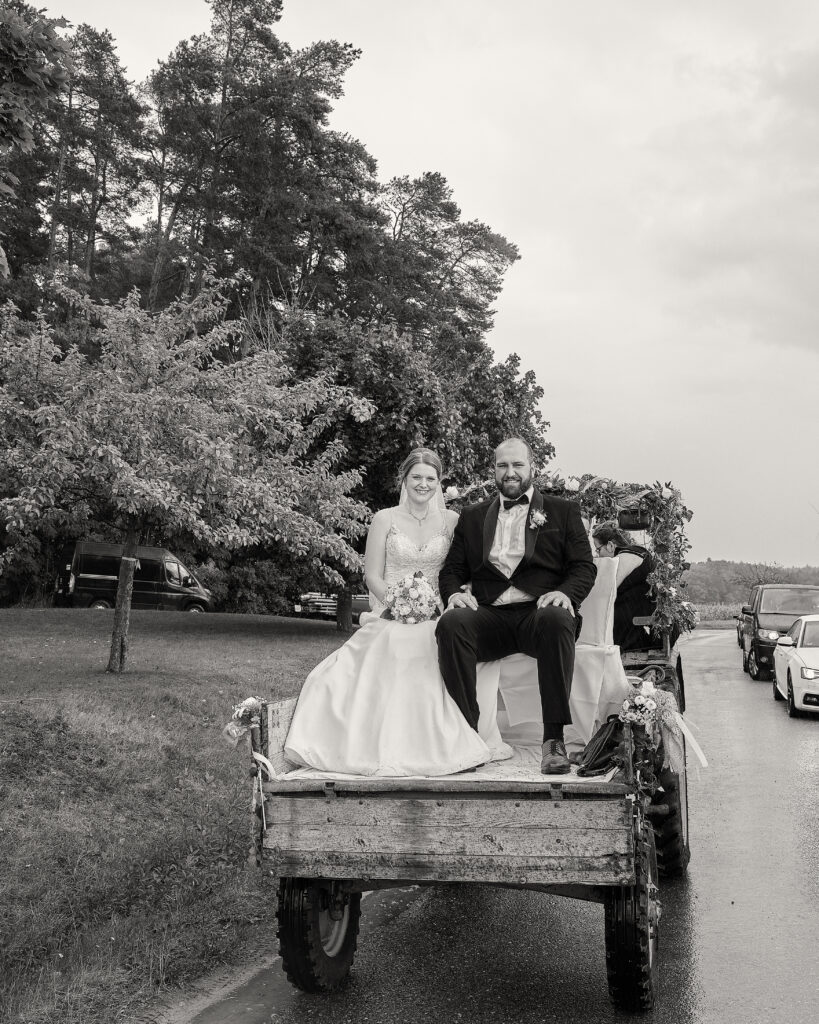 Schwarz-Weiß Bild. Braut und Bräutigam sitzen auf dem Schaufellader des geschmückten Traktors und schauen in die Kamera.