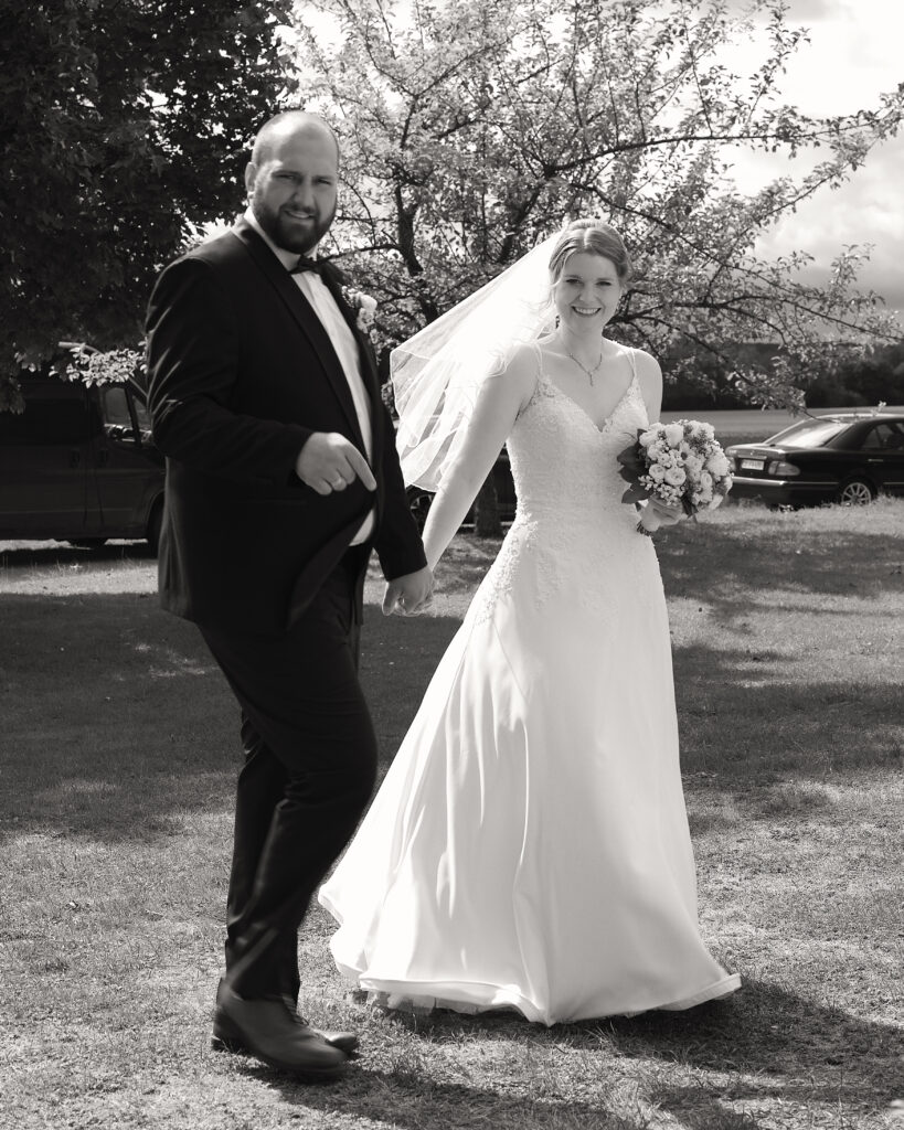 Schwarz-Weiß Bild. Braut und Bräutigam draußen als Ganzkörper Bild schauen in die Kamera.