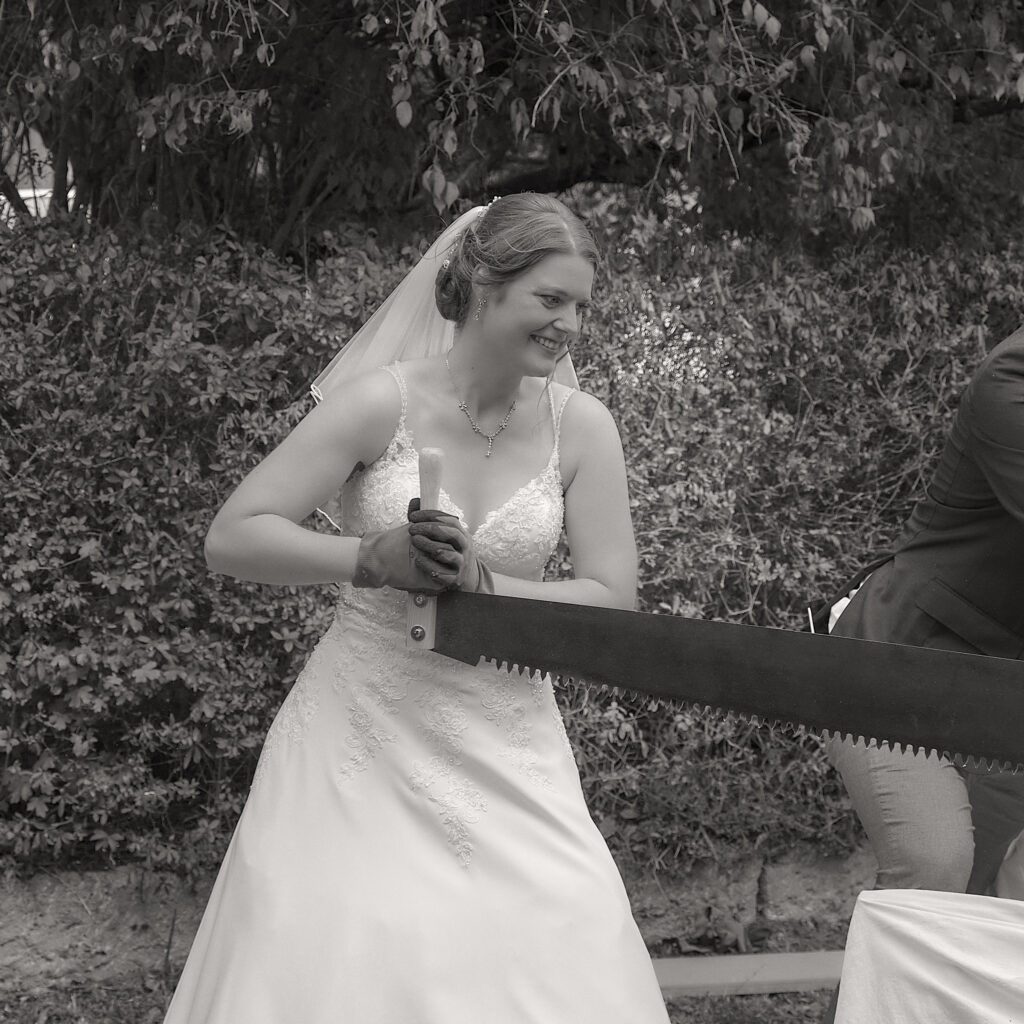 Schwarz-Weiß Hochzeitsfoto. Braut sägt zum Hochzeitsbrauch ein Holz durch.
