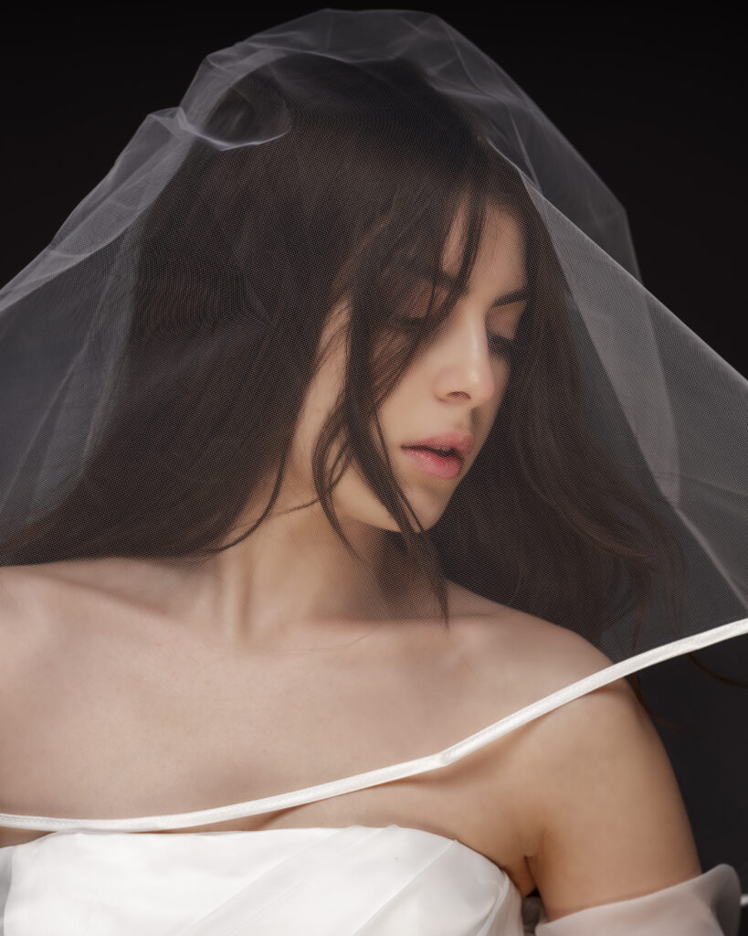 Eine junge Braut in engem Bildausschnitt und mit Brautschleier über dem Kopf. Hintergrund schwarz