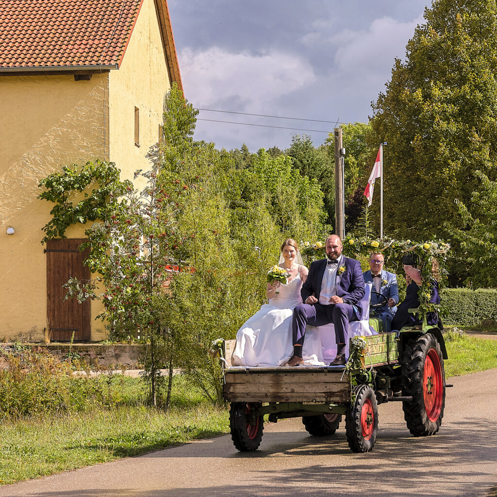 Ein Traktor mit vorne liegender Ladefläche, auf der das Brautpaar sitzt. Er ist als Hochzeitsauto geschmückt und fährt durchs Dorf.