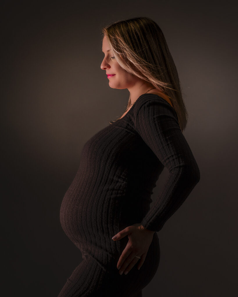 Ein dunkles Bild einer hochschwangeren blonden Frau im Profil bis zum Oberschenkel mit braunem Hintergrund.
